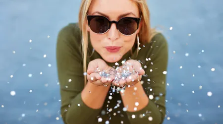 Woman blowing glitter towards camera_small_shutterstock Close up portrait of attractive young woman blowing glitters. Caucasian Female model having fun over blue background.