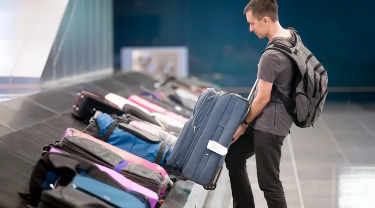 Young handsome man passenger in 20s with carry-on backpack collecting his luggage at conveyor belt in arrivals lounge of airport terminal building