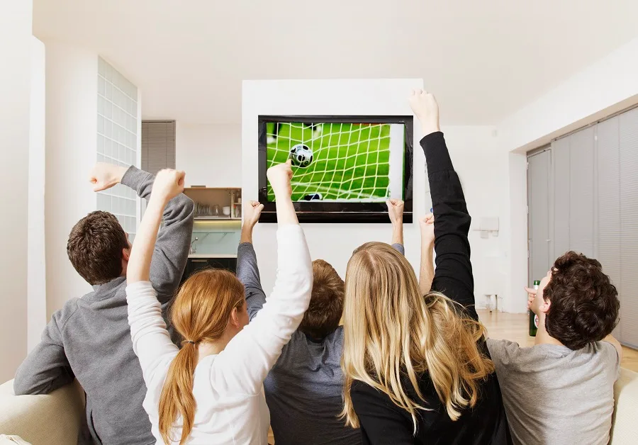 Image: Getty Images Four adults cheering at soccer playing on TV.