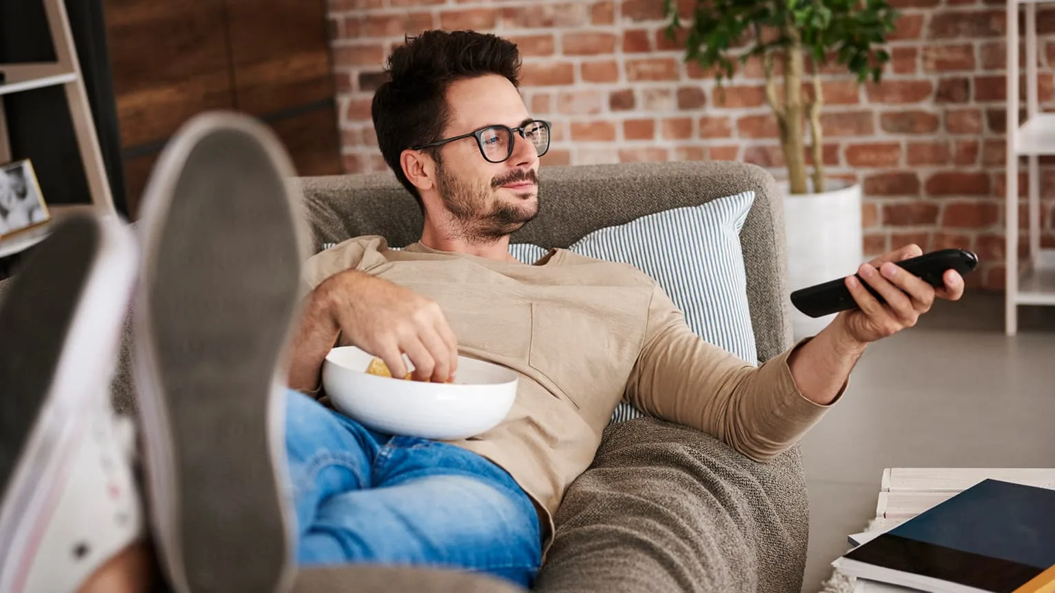 Image: Getty Images Content man lying on couch at home with bowl of potato chips watching TV