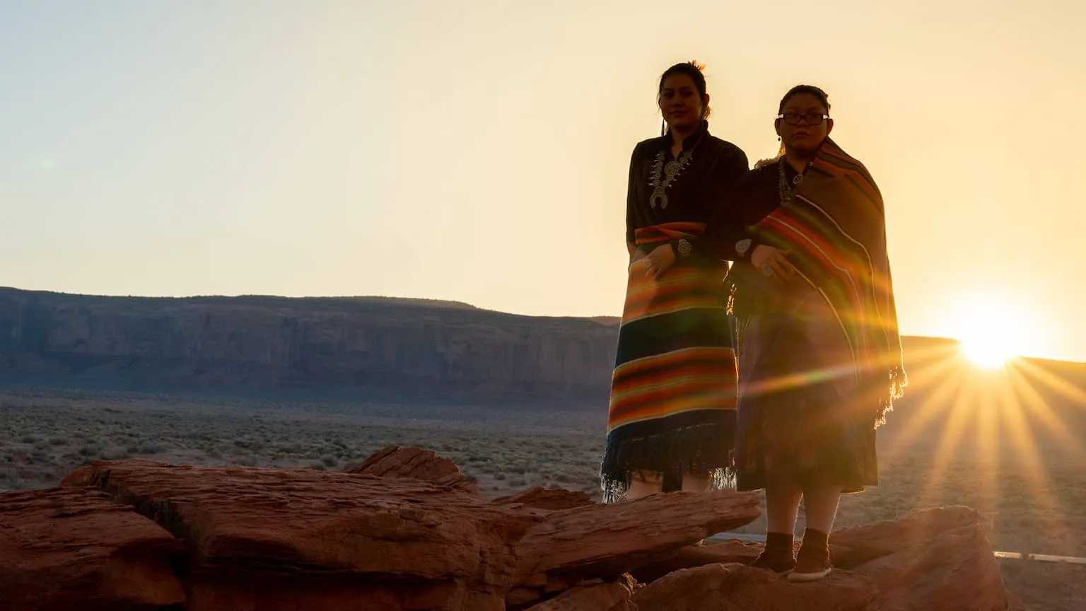 Image: Getty Images Two Indigenous women standing on a mesa with a sunset in the background.