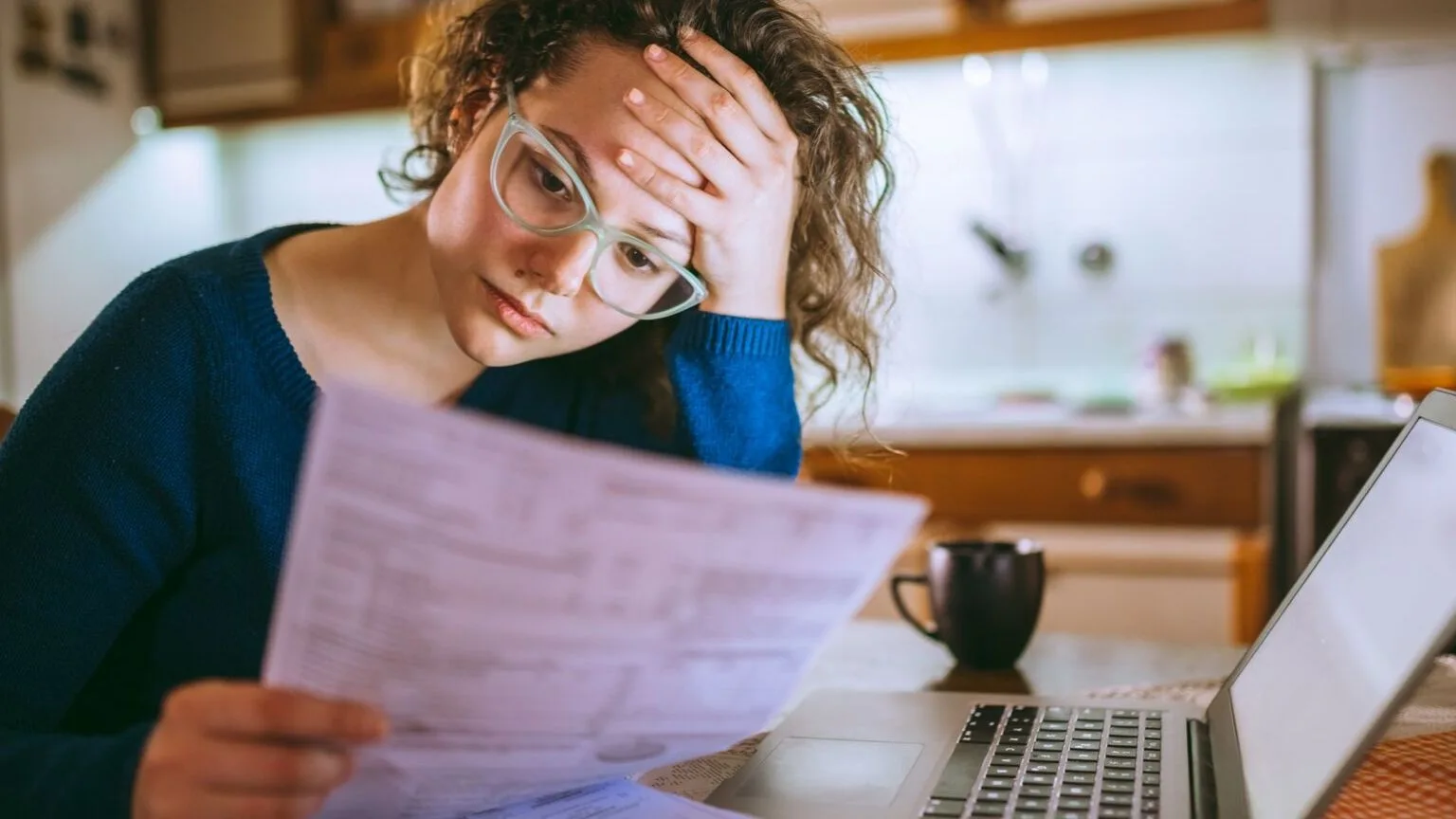 Image: Getty Images Stressed woman reading bills or paperwork in front of laptop.