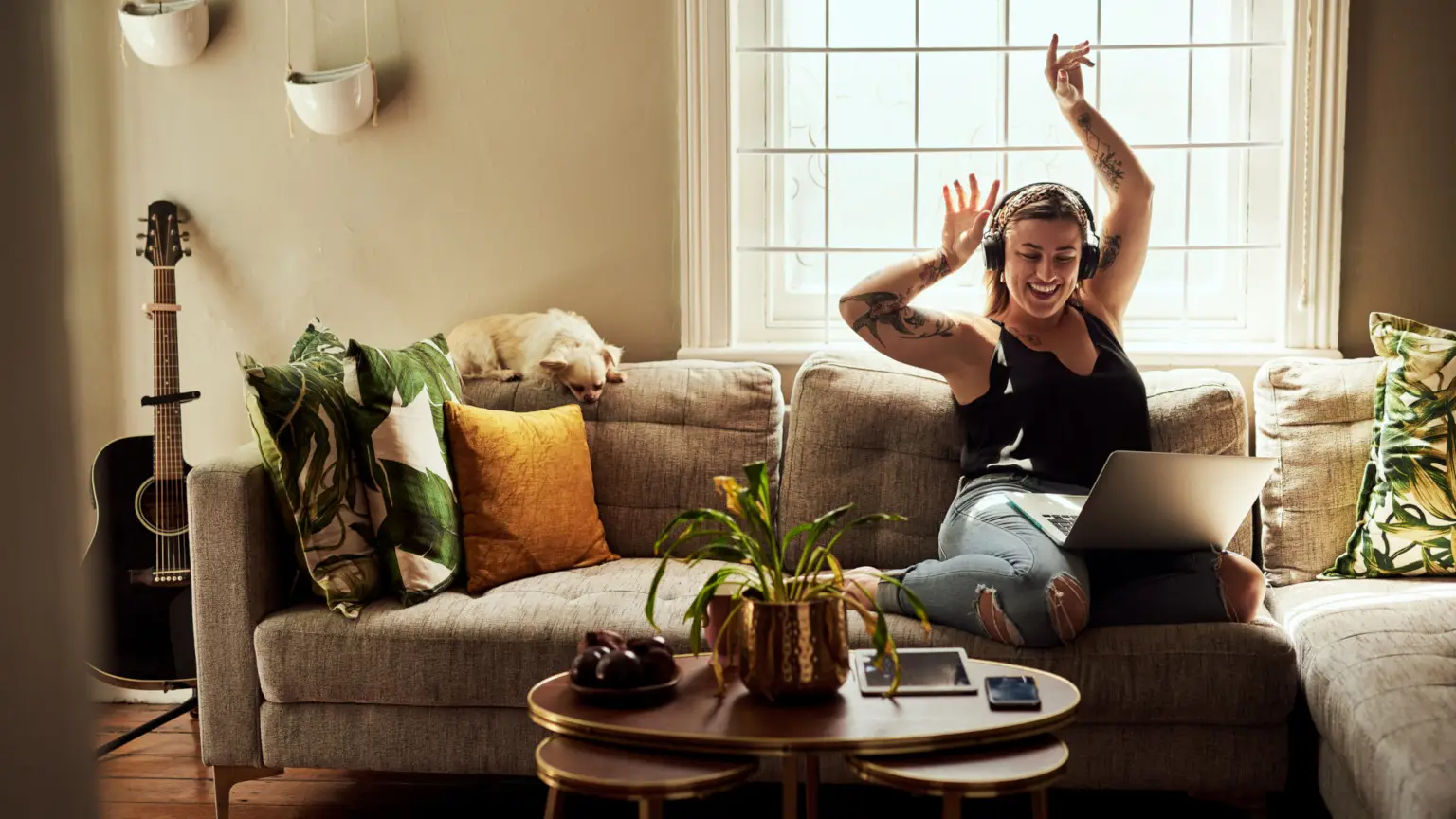 Image: Getty Images Woman sitting on her couch with laptop on headphones with her hands in the air