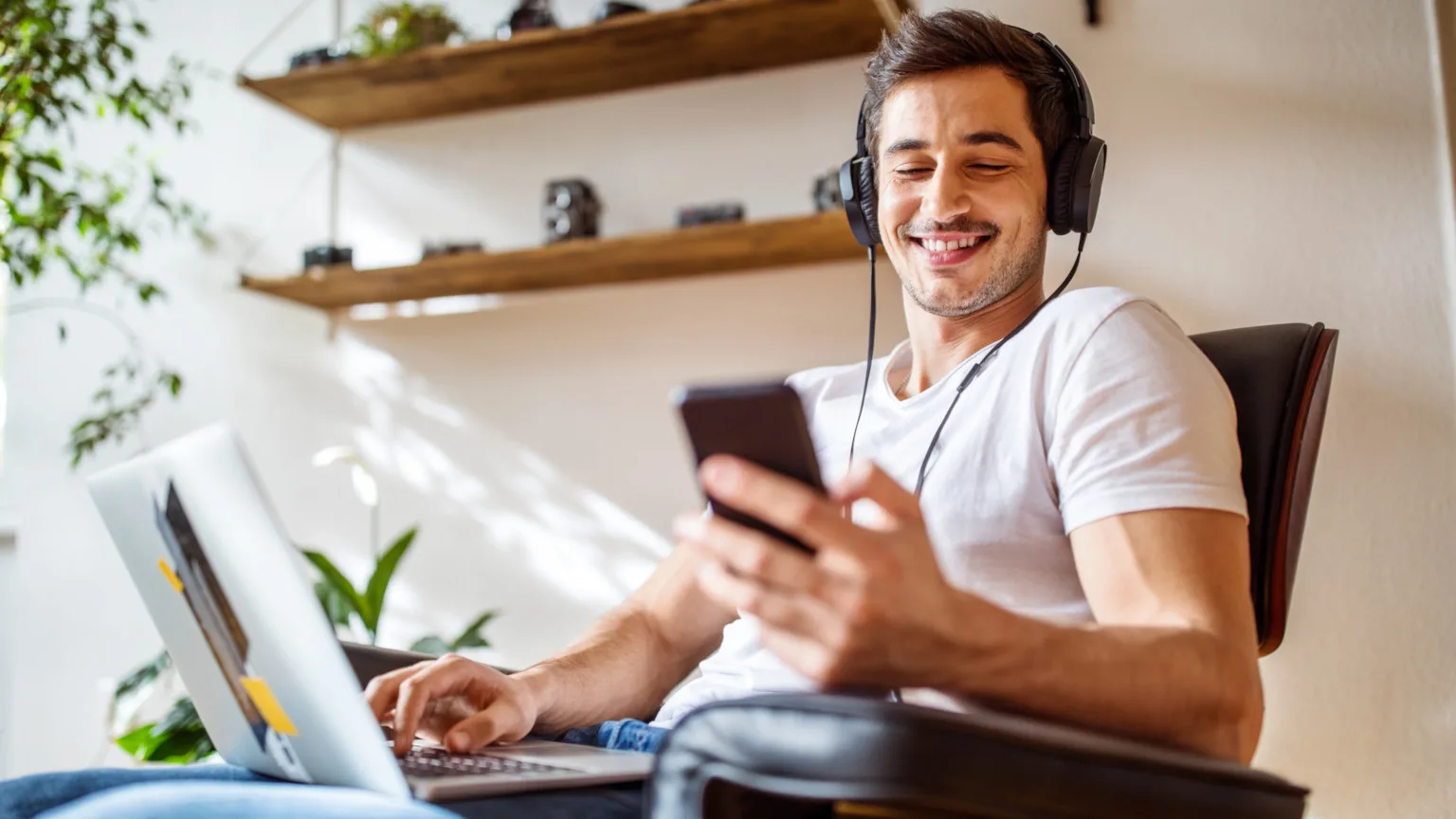 Young man wearing headphones looking at his smart phone with a laptop on his lap
