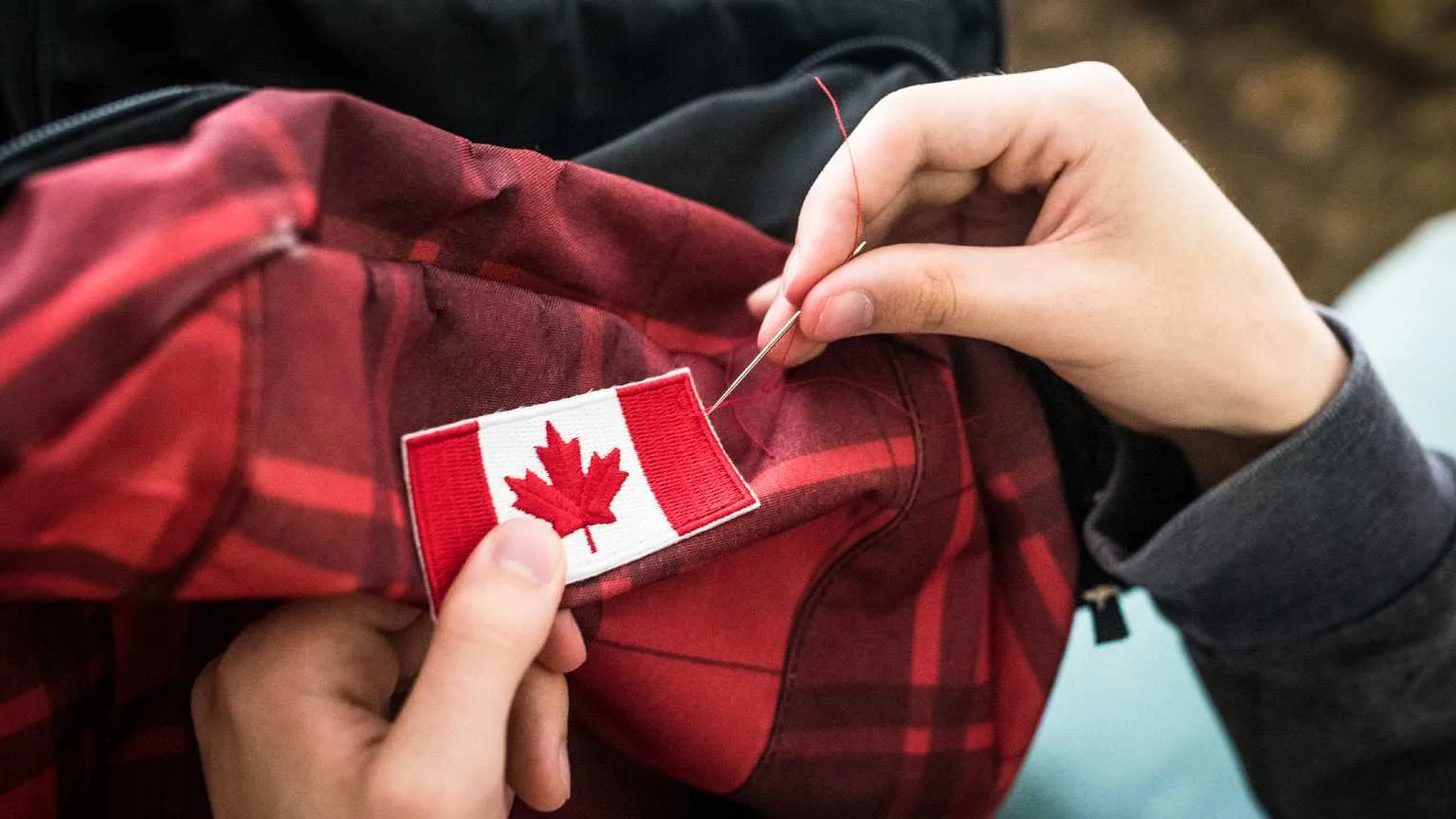 Image: Getty Images Close up personal perspective of a young adult's hands sewing a maple leaf Canada flag patch onto a backpack for travel.