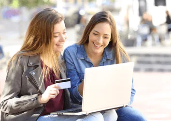 Two friends checking a basic savings account a laptop sitting on a bench